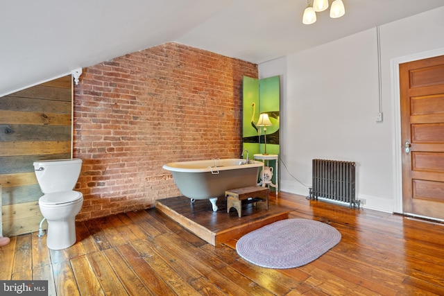 bathroom featuring brick wall, a bath, wood-type flooring, radiator heating unit, and toilet