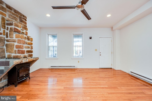 unfurnished living room with a baseboard heating unit, light wood-type flooring, a wood stove, and ceiling fan