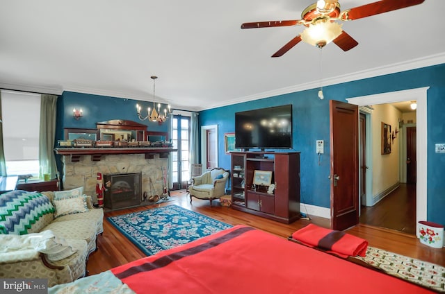 living room featuring wood finished floors, ceiling fan with notable chandelier, a stone fireplace, baseboards, and crown molding