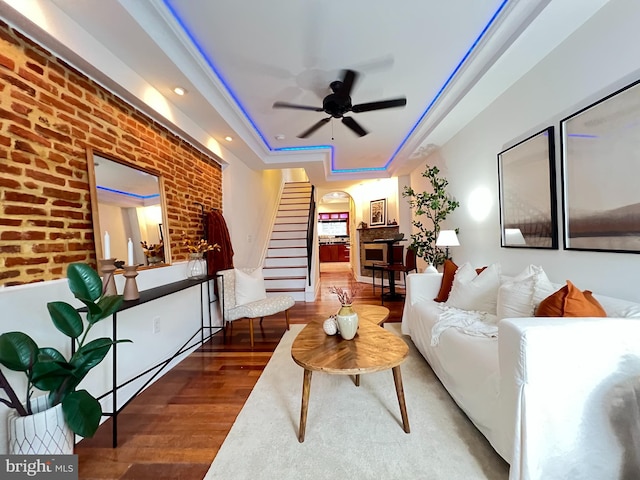 living room featuring wood finished floors, a tray ceiling, brick wall, stairway, and ceiling fan