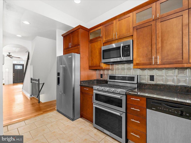 kitchen with brown cabinetry, dark stone counters, light tile patterned flooring, decorative backsplash, and appliances with stainless steel finishes