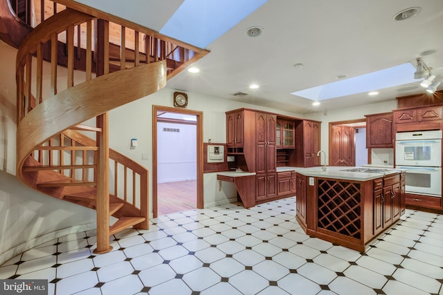 kitchen with a kitchen island with sink, sink, a skylight, stainless steel gas cooktop, and white double oven