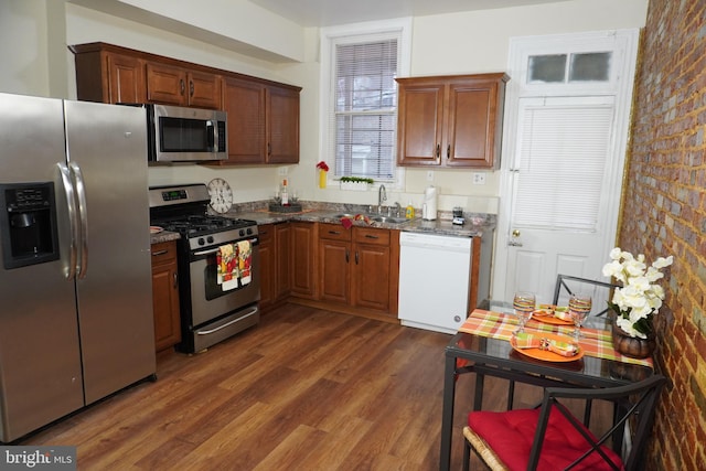 kitchen featuring dark stone countertops, brick wall, sink, appliances with stainless steel finishes, and dark hardwood / wood-style flooring