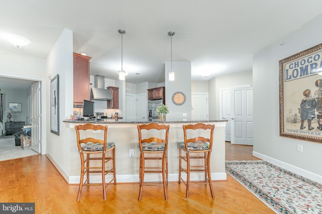 kitchen featuring kitchen peninsula, wall chimney exhaust hood, hanging light fixtures, stainless steel fridge, and light wood-type flooring