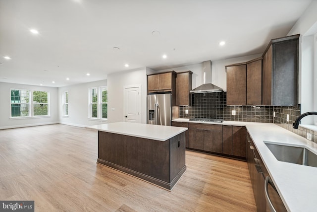 kitchen with wall chimney range hood, appliances with stainless steel finishes, light wood-type flooring, sink, and a center island