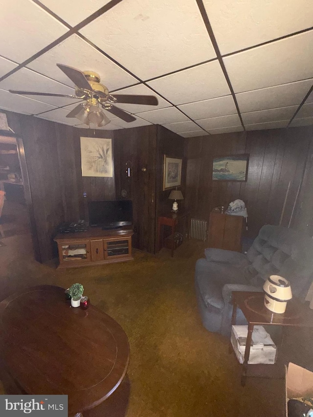 carpeted living room featuring radiator, ceiling fan, wood walls, and a drop ceiling