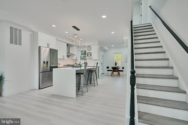 kitchen featuring wall chimney range hood, stainless steel fridge, white cabinetry, light hardwood / wood-style floors, and pendant lighting