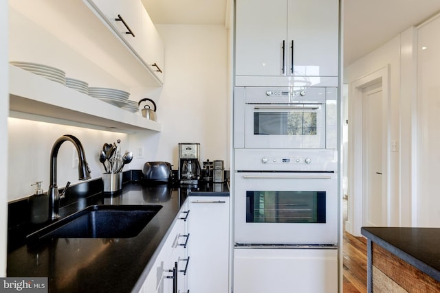 kitchen featuring white double oven, a sink, white cabinets, open shelves, and dark countertops