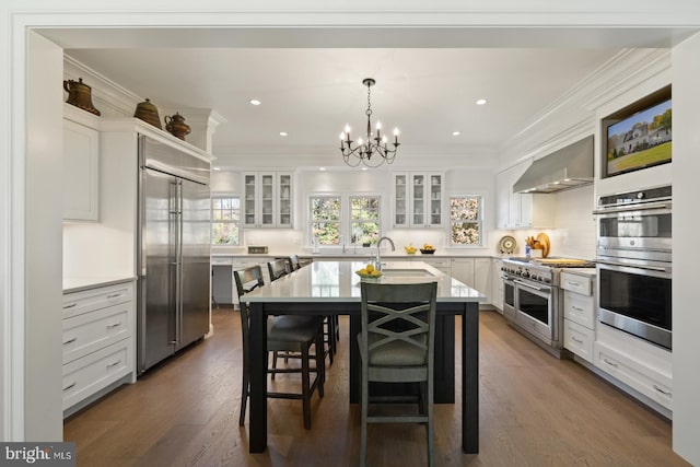 kitchen featuring wall chimney range hood, white cabinets, dark wood-type flooring, high end appliances, and a center island with sink