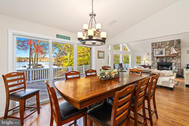 dining room with a stone fireplace, vaulted ceiling, a notable chandelier, light wood-type flooring, and a water view