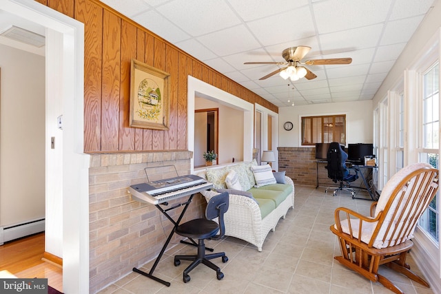 living room with light tile patterned floors, ceiling fan, wood walls, a drop ceiling, and brick wall