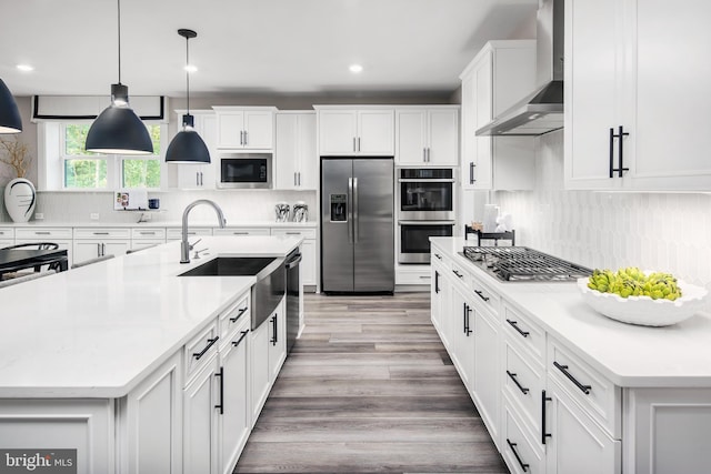 kitchen featuring wall chimney range hood, stainless steel appliances, pendant lighting, light hardwood / wood-style flooring, and a kitchen island with sink