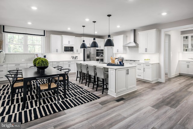 kitchen featuring wall chimney range hood, an island with sink, appliances with stainless steel finishes, white cabinetry, and light hardwood / wood-style floors