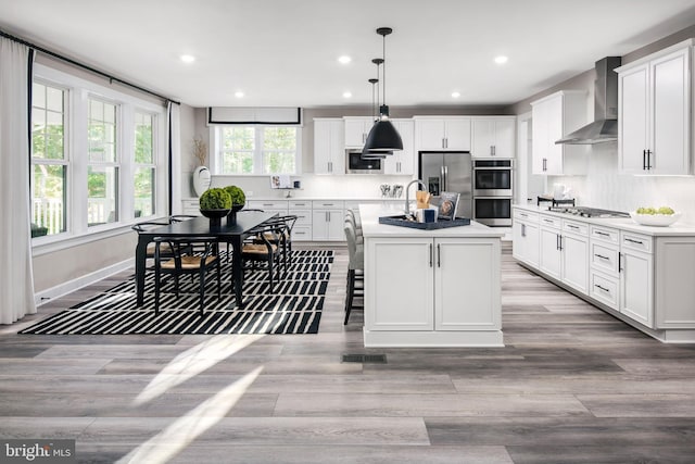 kitchen featuring a center island with sink, white cabinets, wall chimney range hood, and decorative light fixtures