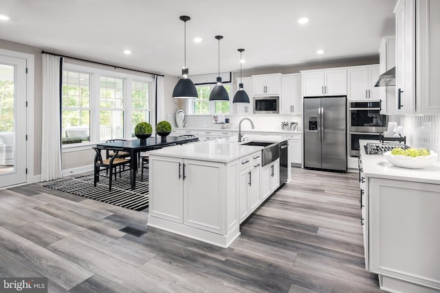 kitchen featuring an island with sink, stainless steel appliances, pendant lighting, light wood-type flooring, and white cabinets