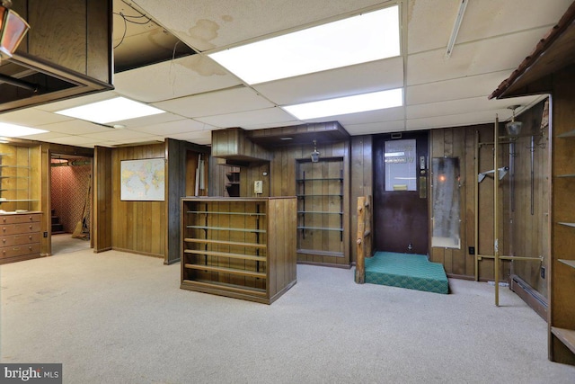 basement featuring light colored carpet, a paneled ceiling, and wooden walls