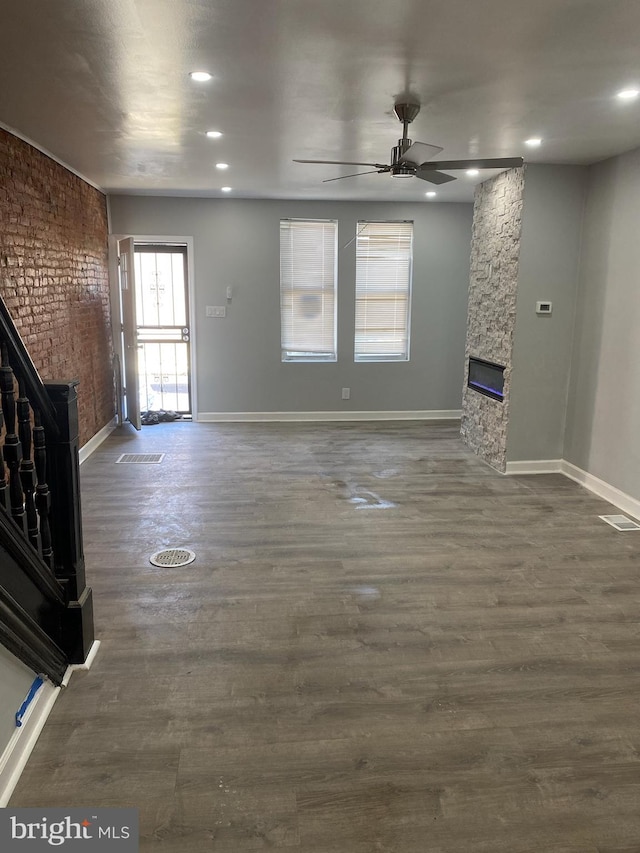 unfurnished living room with brick wall, dark wood-type flooring, a stone fireplace, and ceiling fan