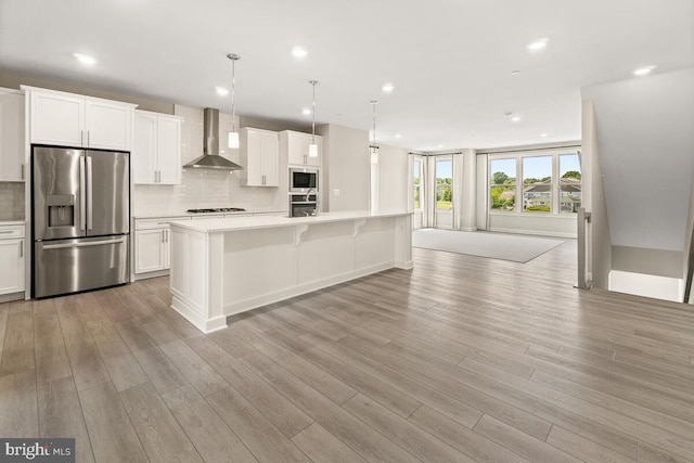 kitchen featuring wall chimney range hood, light hardwood / wood-style floors, stainless steel appliances, decorative light fixtures, and a kitchen island with sink