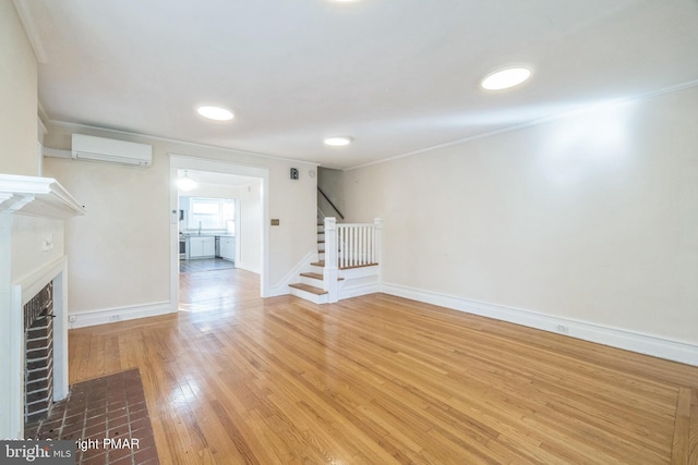 unfurnished living room with a brick fireplace, stairs, baseboards, an AC wall unit, and wood finished floors
