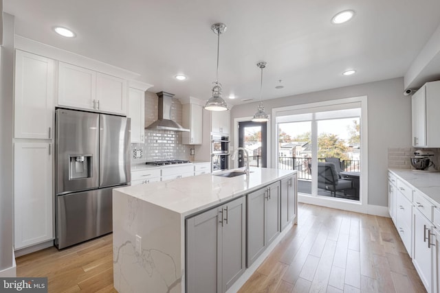 kitchen with appliances with stainless steel finishes, white cabinetry, a kitchen island with sink, wall chimney exhaust hood, and light stone counters