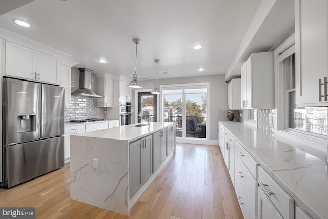 kitchen featuring a kitchen island with sink, wall chimney range hood, white cabinets, pendant lighting, and appliances with stainless steel finishes