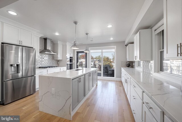 kitchen featuring wall chimney range hood, stainless steel appliances, decorative light fixtures, white cabinets, and a center island with sink