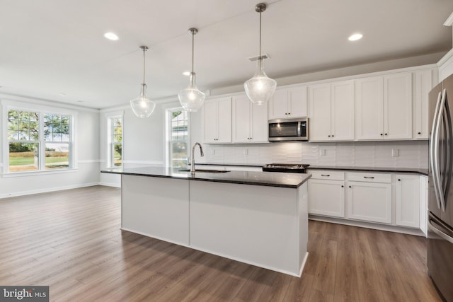 kitchen with an island with sink, appliances with stainless steel finishes, dark wood-type flooring, sink, and decorative light fixtures