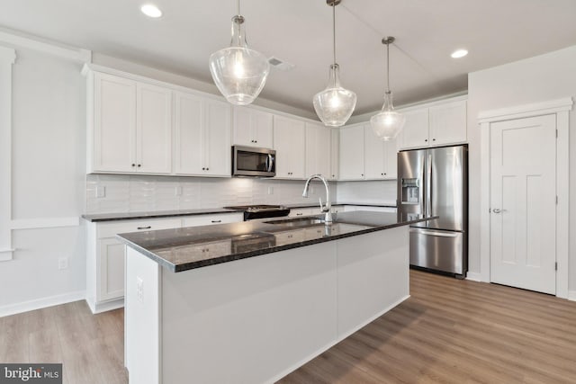 kitchen with an island with sink, stainless steel appliances, decorative light fixtures, white cabinetry, and light hardwood / wood-style floors