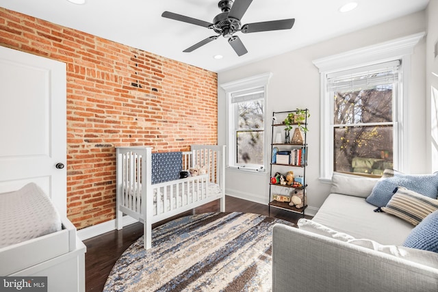 bedroom with a crib, ceiling fan, brick wall, and dark hardwood / wood-style flooring