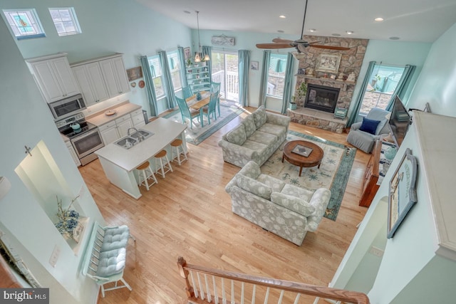 living room with a stone fireplace, ceiling fan with notable chandelier, sink, high vaulted ceiling, and light hardwood / wood-style flooring