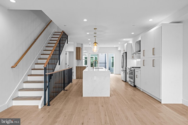 kitchen with light wood finished floors, appliances with stainless steel finishes, a kitchen island with sink, a sink, and wall chimney range hood