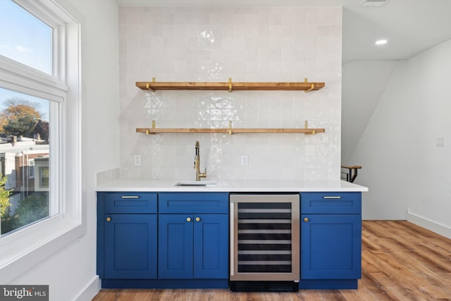 bar featuring wood-type flooring, beverage cooler, blue cabinets, decorative backsplash, and sink