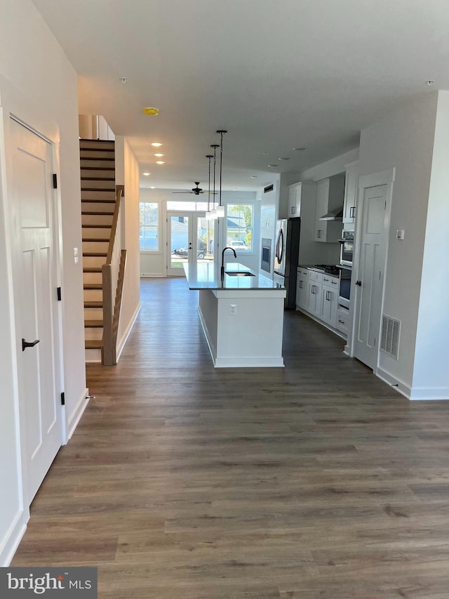 kitchen with appliances with stainless steel finishes, wall chimney exhaust hood, dark wood-type flooring, decorative light fixtures, and an island with sink