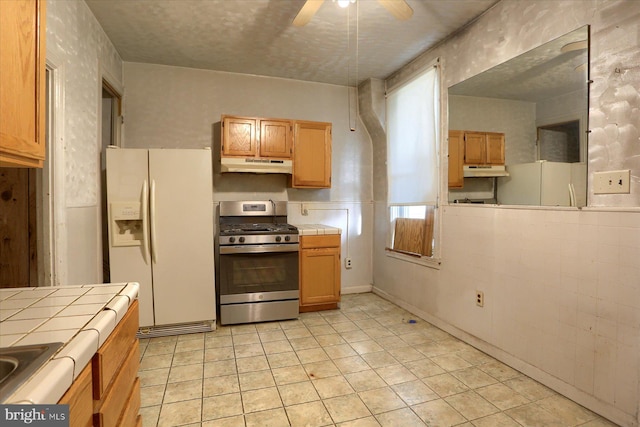 kitchen featuring white fridge with ice dispenser, light tile patterned flooring, stainless steel range with gas stovetop, ceiling fan, and tile counters