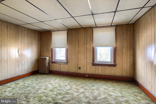 carpeted empty room with radiator, a healthy amount of sunlight, a drop ceiling, and wood walls