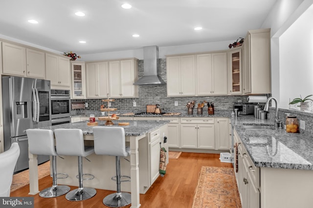 kitchen with light stone counters, wall chimney exhaust hood, stainless steel appliances, a center island, and light hardwood / wood-style floors