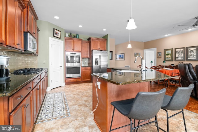 kitchen featuring decorative backsplash, hanging light fixtures, stainless steel appliances, dark stone counters, and a center island with sink