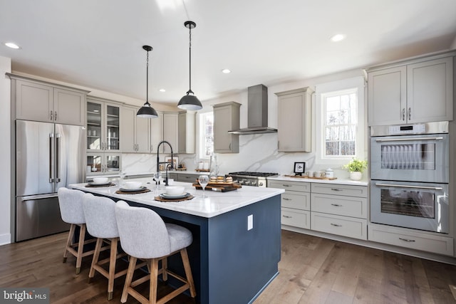 kitchen with wall chimney range hood, appliances with stainless steel finishes, dark hardwood / wood-style flooring, hanging light fixtures, and a kitchen island with sink