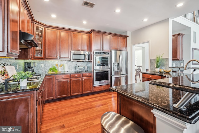 kitchen featuring decorative backsplash, appliances with stainless steel finishes, light wood-type flooring, dark stone counters, and extractor fan