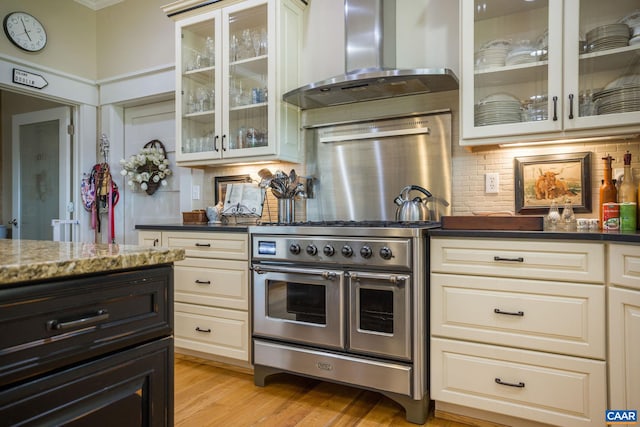 kitchen featuring wall chimney range hood, range with two ovens, dark stone counters, decorative backsplash, and light hardwood / wood-style flooring