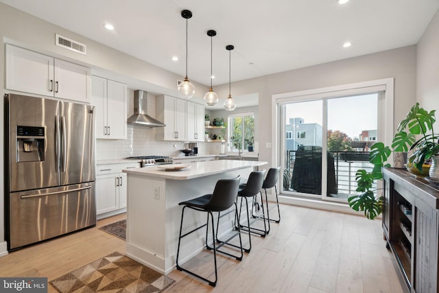 kitchen with wall chimney range hood, appliances with stainless steel finishes, white cabinetry, and hanging light fixtures