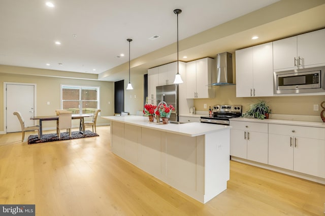 kitchen with appliances with stainless steel finishes, wall chimney range hood, a center island with sink, decorative light fixtures, and light hardwood / wood-style floors