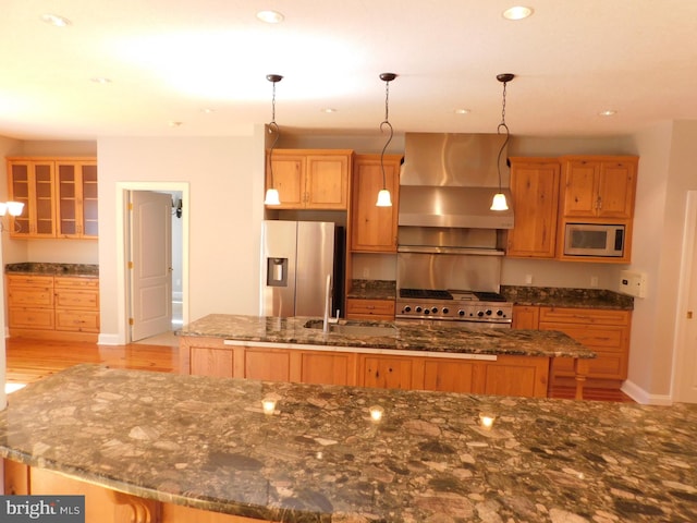 kitchen featuring wall chimney range hood, appliances with stainless steel finishes, an island with sink, and hanging light fixtures