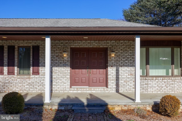 view of exterior entry with roof with shingles, a porch, and brick siding