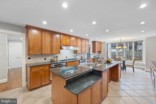 kitchen featuring wall oven, brown cabinetry, a kitchen island with sink, under cabinet range hood, and stainless steel gas stovetop