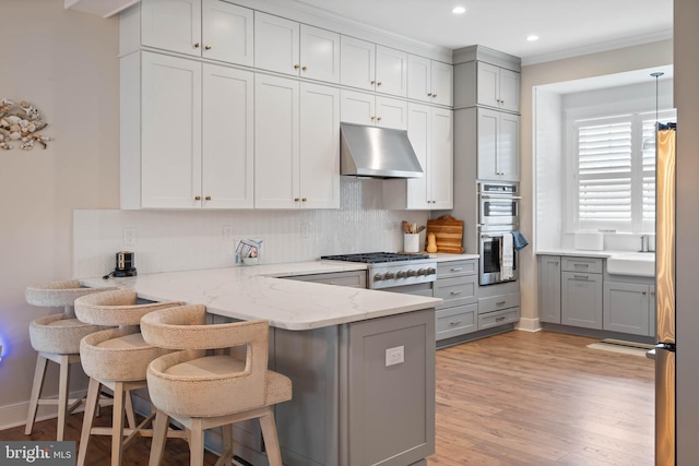 kitchen with appliances with stainless steel finishes, kitchen peninsula, a kitchen bar, range hood, and light wood-type flooring