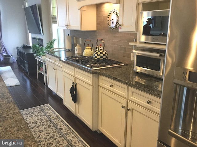 kitchen featuring appliances with stainless steel finishes, cream cabinets, dark wood-type flooring, dark stone countertops, and decorative backsplash