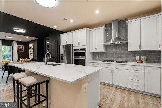 kitchen featuring wall chimney exhaust hood, appliances with stainless steel finishes, an island with sink, and white cabinets