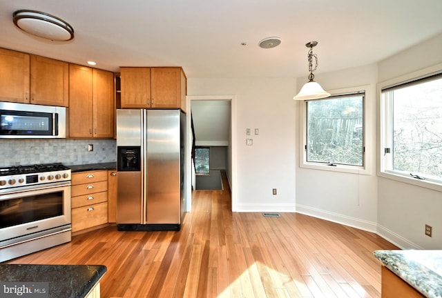 kitchen with light wood-type flooring, backsplash, dark stone counters, stainless steel appliances, and hanging light fixtures