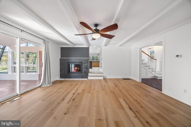 unfurnished living room featuring beam ceiling, a stone fireplace, light wood-type flooring, and ceiling fan